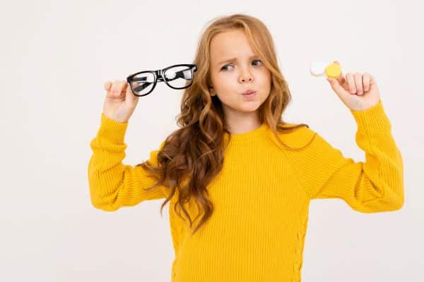 Menina segurando um par de óculos de grau numa mão e noutra um estojo de lentes de contato. Qual a melhor escolha para crianças?