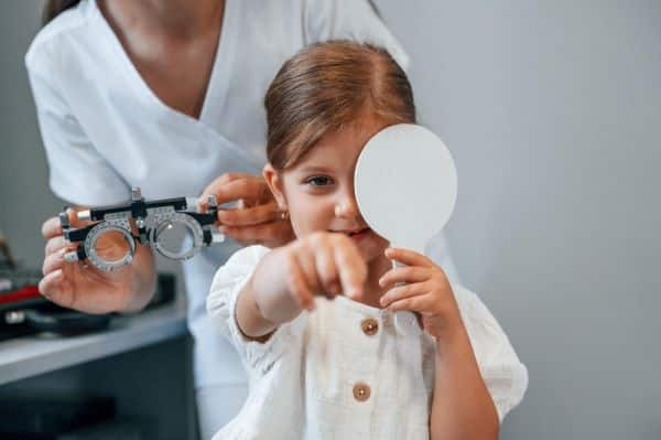 Uma garotinha realizando exame oftalmológico para identificar erros refrativos. Ela cobre um dos olhos enquanto aponta com o dedo para algo à sua frente.