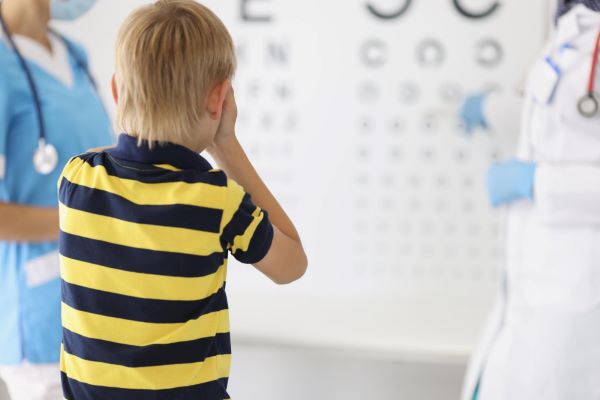 Erros refrativos: garotinho loiro realizando consulta oftalmológica. Ele tenta ler as letras de um painel de teste para acuidade visual.