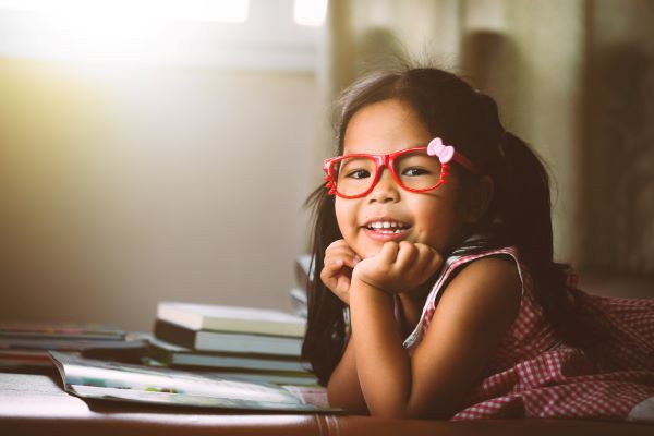 Menina asiática, debruçada sobre livros, usando um óculos vermelho.