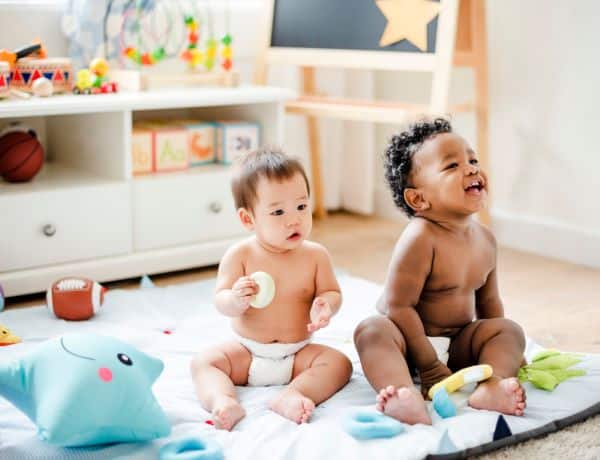 Dois bebês de etnias distintas, um oriental e outro negro, estão sentados num tapete no quarto brincando. 
