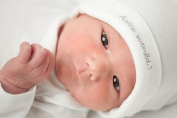 Bebê recém-nascido deitado em fundo branco, usa toquinha branca na qual está escrito:" Olá Mundo!". 
Conceito de catarata congênita