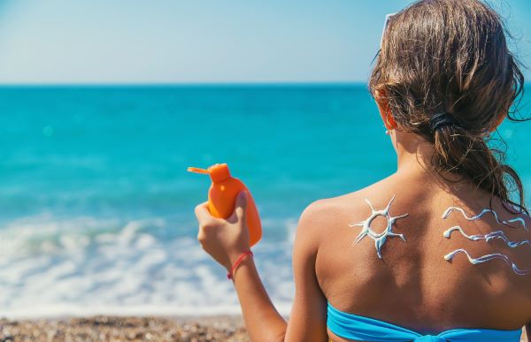 Menina frente ao mar, segura um tubo de protetor solar e tem, em suas costas o desenho de um sol e ondas feitos com o creme do protetor.