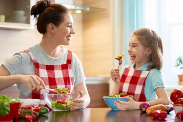 Mãe e filha pequena, felizes e com aventais xadrezes em vermelho e branco, preparam uma salada saudável. Conceito de alimentação equilibrada.