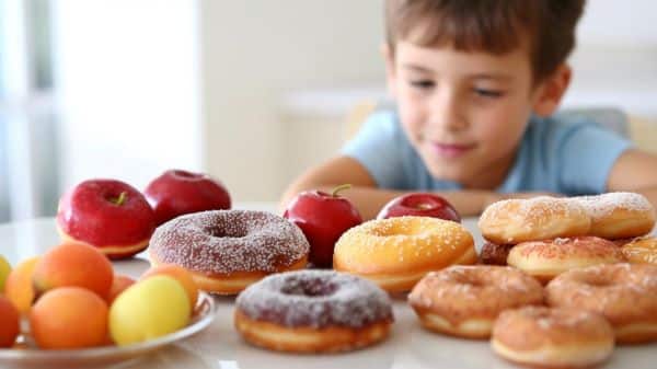 Menino frente a uma mesa, na qual estão frutas e rosquinhas doces, vive o dilema da escolha alimentar.  Alimentação saudável é essencial na infância.
