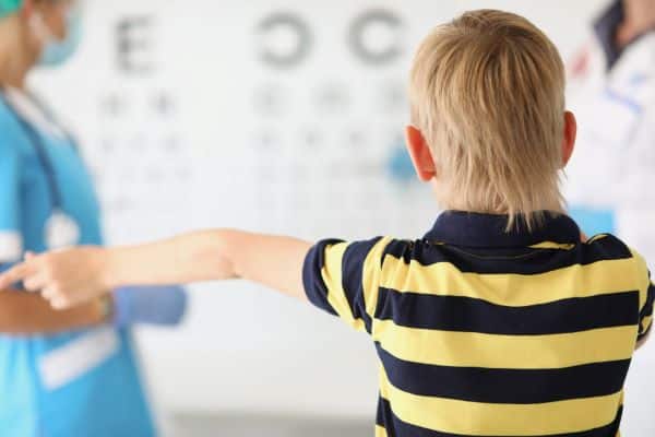 Garotinho loiro, vestindo uma camisa listrada de azul e amarelo, realiza exame ocular no consultório do oftalmologista.