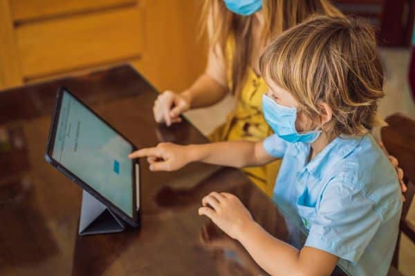 Menino, vestindo uma camisa azul clara, realiza os temas de casa em um tablet com a ajuda materna. Ambos estão usando máscaras em alusão ao período da pandemia.