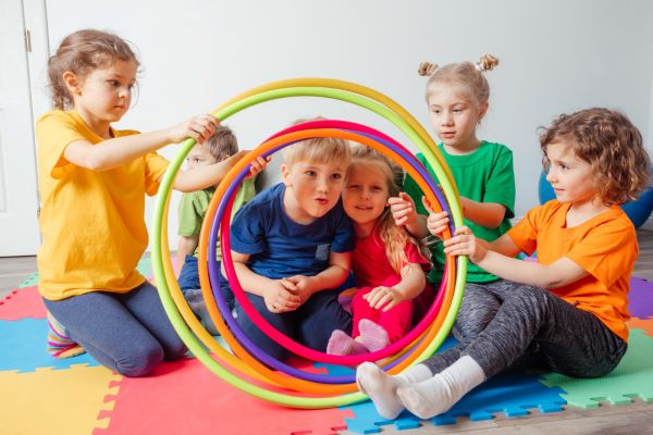 Grupo de crianças com cerca de sete anos, sentadas sobre um tapete de EVA colorido, fazem um túnel com bambolês multicoloridos para brincar.
