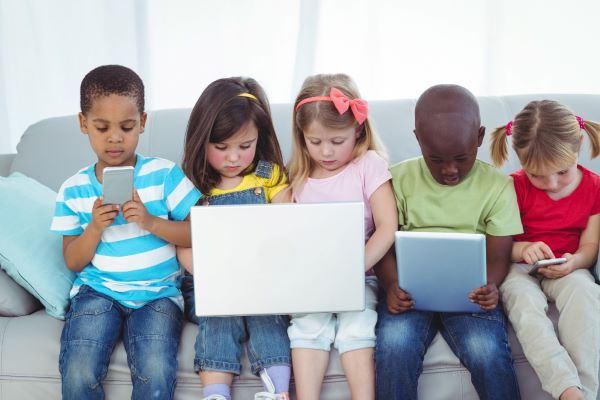 5 crianças de diferentes etnias sentadas num sofá cinza e brincando com tablets e celulares.