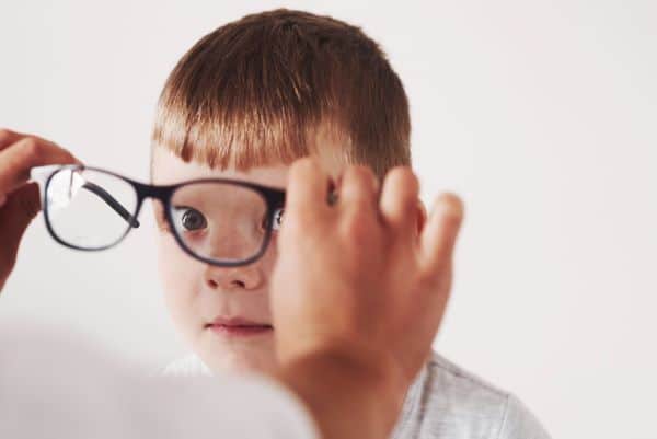 Menino pequeno, loiro com olhos claros, está sendo testado por uma oftalmologista que coloca um par de óculos frente ao seu rostinho para averiguar condições oculares como ambioplia e estrabismo.