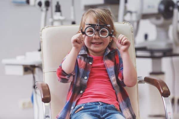 Menininha sorridente, vestindo calça jeans, camiseta rosa e camisa xadrez de rosa e azul por cima, segura um instrumento de testagem oftalmológica frente aos olhos para identificação de doenças como estrabismo e ambioplia.