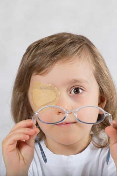 Menino com cerca de 5 anos, com cabelos compridos e loiros. usa um tampão ocular no olho direito e segura um par de óculos de grau azul. 
O uso do tampão ocular é um dos tratamentos tanto do estrabismo como da ambioplia.