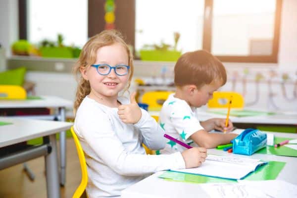 Meninha loira, vestindo camiseta branca e usando um óculos azul claro faz positivo para a foto enquanto realiza tarefa de escola ao lado de um coleguinha loiro.