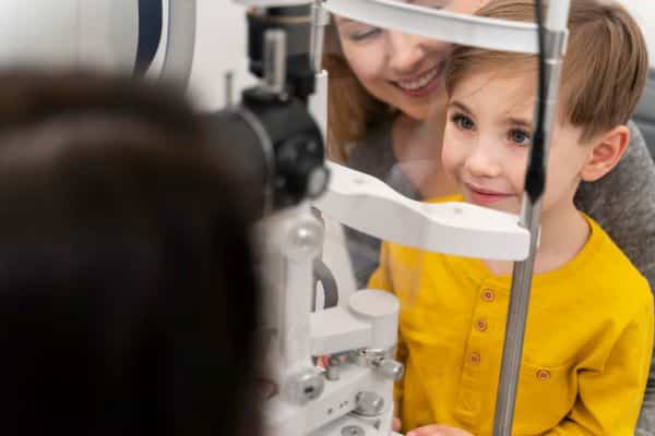 Menininho, sentado no colo de sua mãe, realiza exame ocular para acompanhamento pós-tratamento para retinoblastoma. ele veste uma camiseta amarela.