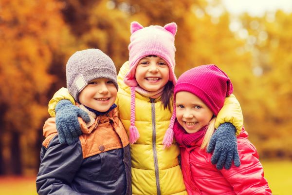 Três crianças, vestindo roupas de inverno coloridas, sorriem abraçadas.