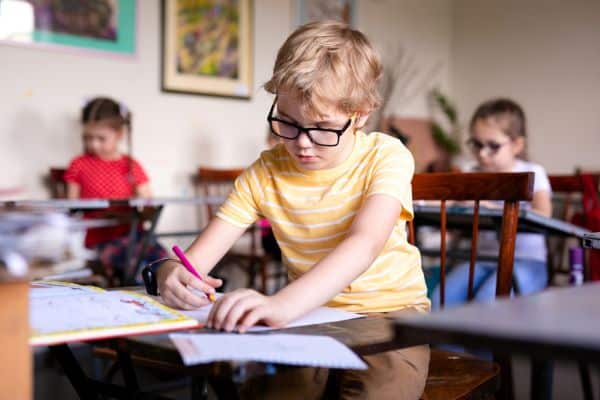 Menino loiro, vestindo camiseta amarela listrada e óculos pretos, realiza atividade em sua classe escolar. Conceito de miopia na infância.