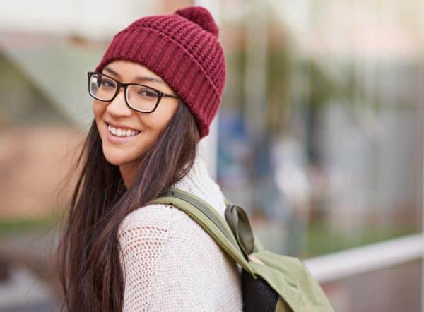 Jovem mulher, com aproximadamente 18 anos, vestindo roupas de inverno e uma mochila verde nas costas, dirige-se à universidade.