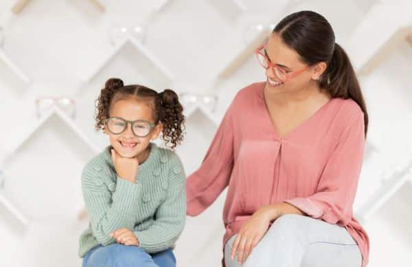 Mãe e filha, usando óculos de grau e vestindo, respectivamente, suéter rosa e suéter verde, sorriem sentadas em um banco.