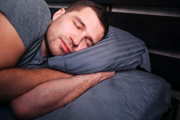 Homem com barba aparente, vestindo camiseta cinza, dorme tranquilamente em sua cama com lençóis cinza.