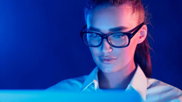 Jovem mulher com cabelos presos em rabo de cavalo, usa um óculos com filtro de luz azul para trabalhar frente ao notebook.