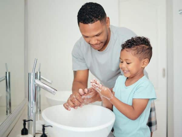 Pai e filho pequeno, negros, lavam juntos as mãos em uma pia branca com torneira prata. Forma de prevenção contra alergias oculares na primavera.