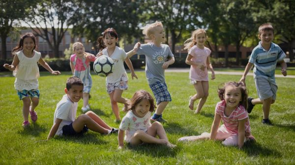 Um grupo de várias crianças em idade escolar divertem-se em um parque ensolarado, jogando futebol.