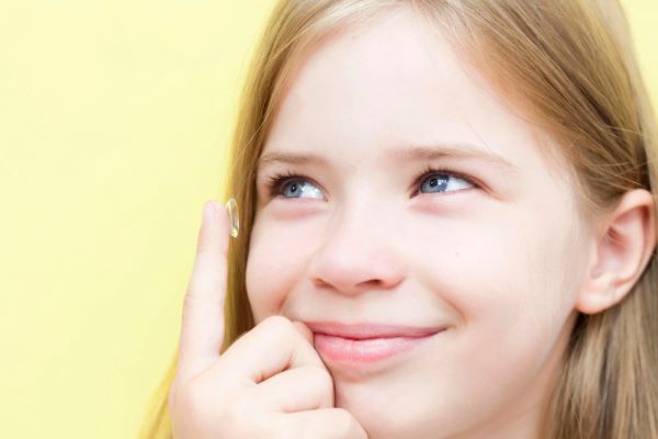 Menina pré-adolescente, loira e com olhos azuis, sorri enquanto coloca sua lente de contato.