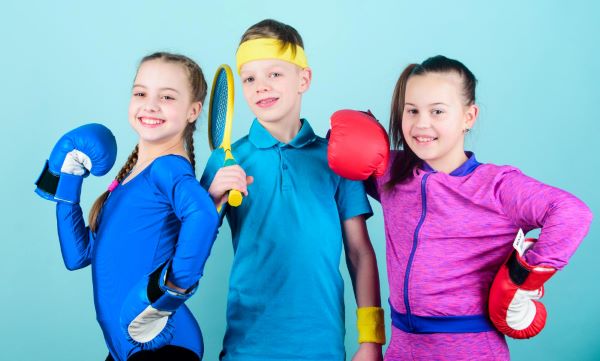 Um menino e duas meninas, usando roupas para realizar atividades físicas e segurando instrumentos de esporte: elas, luvas de box; ele raquete de tênis.