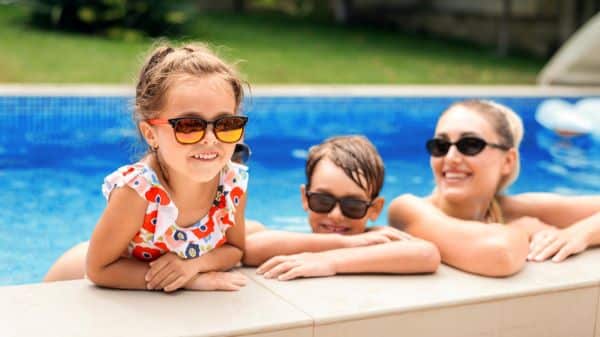 Mãe feliz e dois filhos, um menino maiorzinho e uma menininha, estão apoiados na beira da piscina e protegem-se dos raios uv usando óculos escuros.