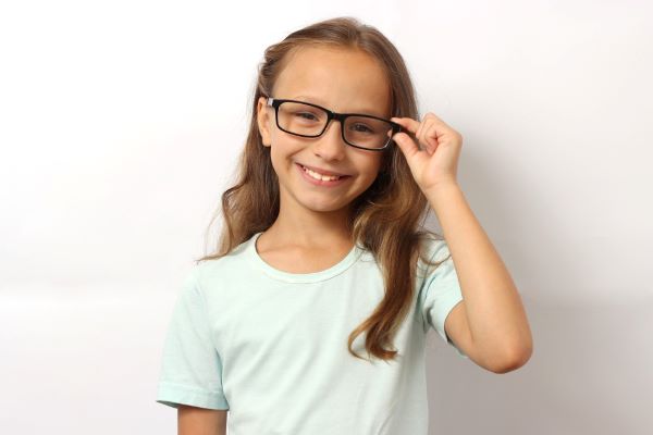 Menina bonita, com cabelos castanhos e longos, vestindo camiseta verde clara, sorri feliz segurando seus óculos. de grau para correção do astigmatismo.
