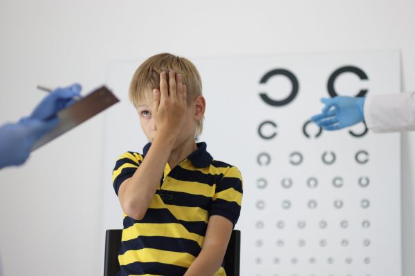 Garoto, em consulta com oftalmologista,  fecha um olho e responde a perguntas.  Está sentado de costas para um painel de testes visuais. 
Ele veste camiseta listrada de preto e amarelo. 