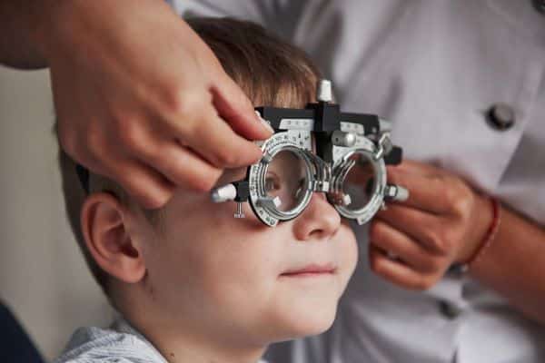 Menininho realiza exame ocular com um instrumento oftalmológico  frente aos olhos. 
As consultas oftalmológicas são essenciais durante a infância. 