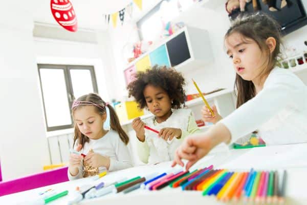 Crianças, frente a uma mesa escolar cheia de lápis de cor e canetinhas coloridas, realizam atividades de pintura .