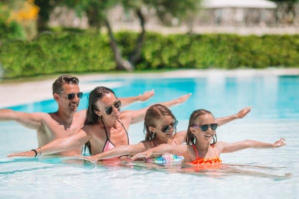 Família composta por pai, mãe e 2 meninas divertem-se na piscina protegendo os olhos com óculos de sol. 