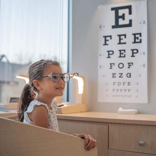 Menina, usando vestido branco com florzinhas e óculos de grau, realiza teste oftalmológico em consultório.