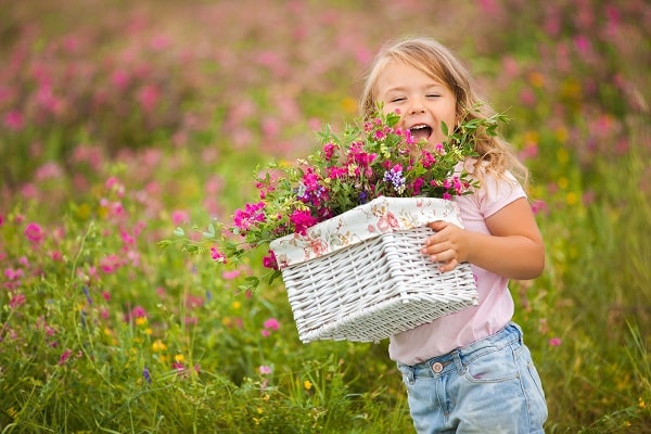 Menina sorridente brinca em meio a um campo coberto de flores coloridas. Ela segura um cesto com flores nas mãos.