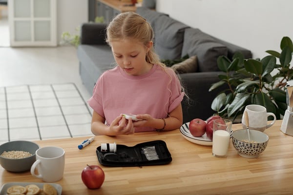 Menina, sentada frente a uma mesa de madeira sobre a qual há prato com maçãs, cerais, copo de leite, 1 caneca e instrumentos de medir a glicose no sangue. A garotinha realiza ela mesma o próprio exame.
