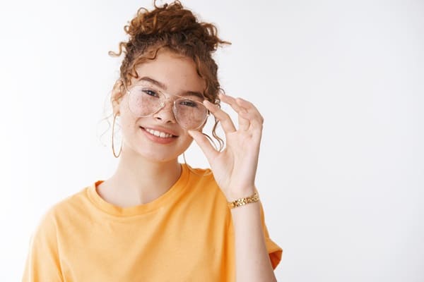 Jovem com cabelos ruivos e cacheados, presos no alto da cabeça, com sardas no rosto, usando óculos de grau e vestindo camiseta amarela, sorri para a câmera.