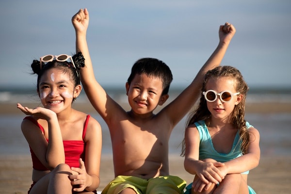Crianças divertindo-se na praia; 2 meninas e 1 menino. Todos com proteção solar.