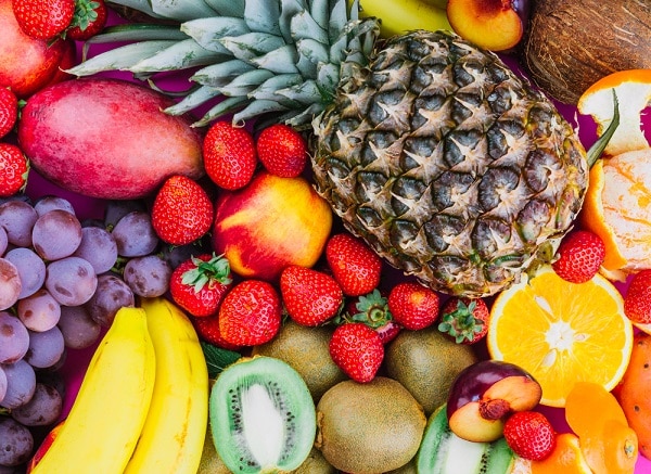 Sobre uma superfície estão frutas como: uvas, morangos, abacaxi, kiwi, damasco, banana, manga e laranja.