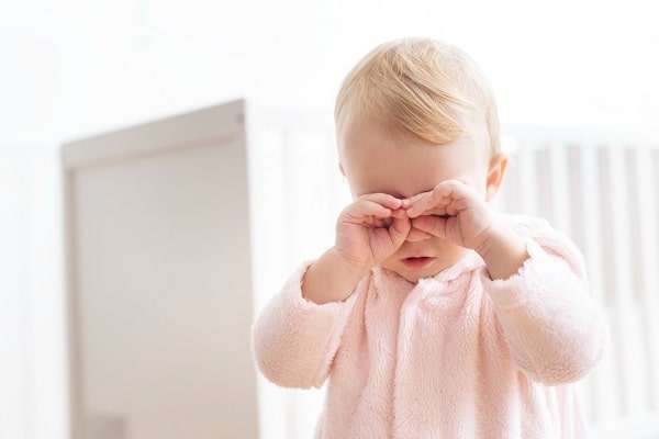 Menininha com cerca de 1 ano de idade, loirinha, vestindo roupa peludinha rosa, coça os olhinhos com as mãos.