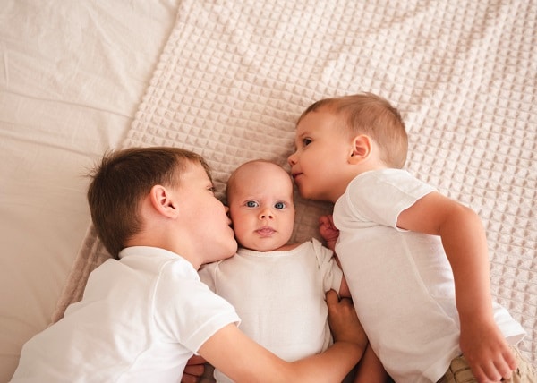Irmãos beijando bebê recém-nascido deitados em uma cama de casal.
