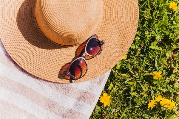 Uma das formas de prevenção do pterígio é o uso constante de chapéu e óculos de sol. Na imagem, um chapéu de palha natural, um par de óculos e uma toalha estão sobre um gramado.