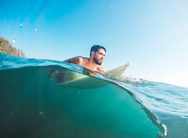 Surfista barbudo deitado em uma prancha de surf sobre o mar.