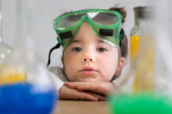 Menina pequena, com os cabelos presos no alto da cabeça, apoia o rosto com as mãos sobre uma bancada de ciências onde estão vários produtos químicos. Para proteger seus olhos de traumas oculares, ela usa um óculos de proteção verde.
