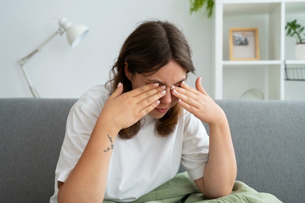 Jovem com cabelos escuros, sentada no sofá da sala, esfrega os olhos demonstrando sinais de desconforto visual.